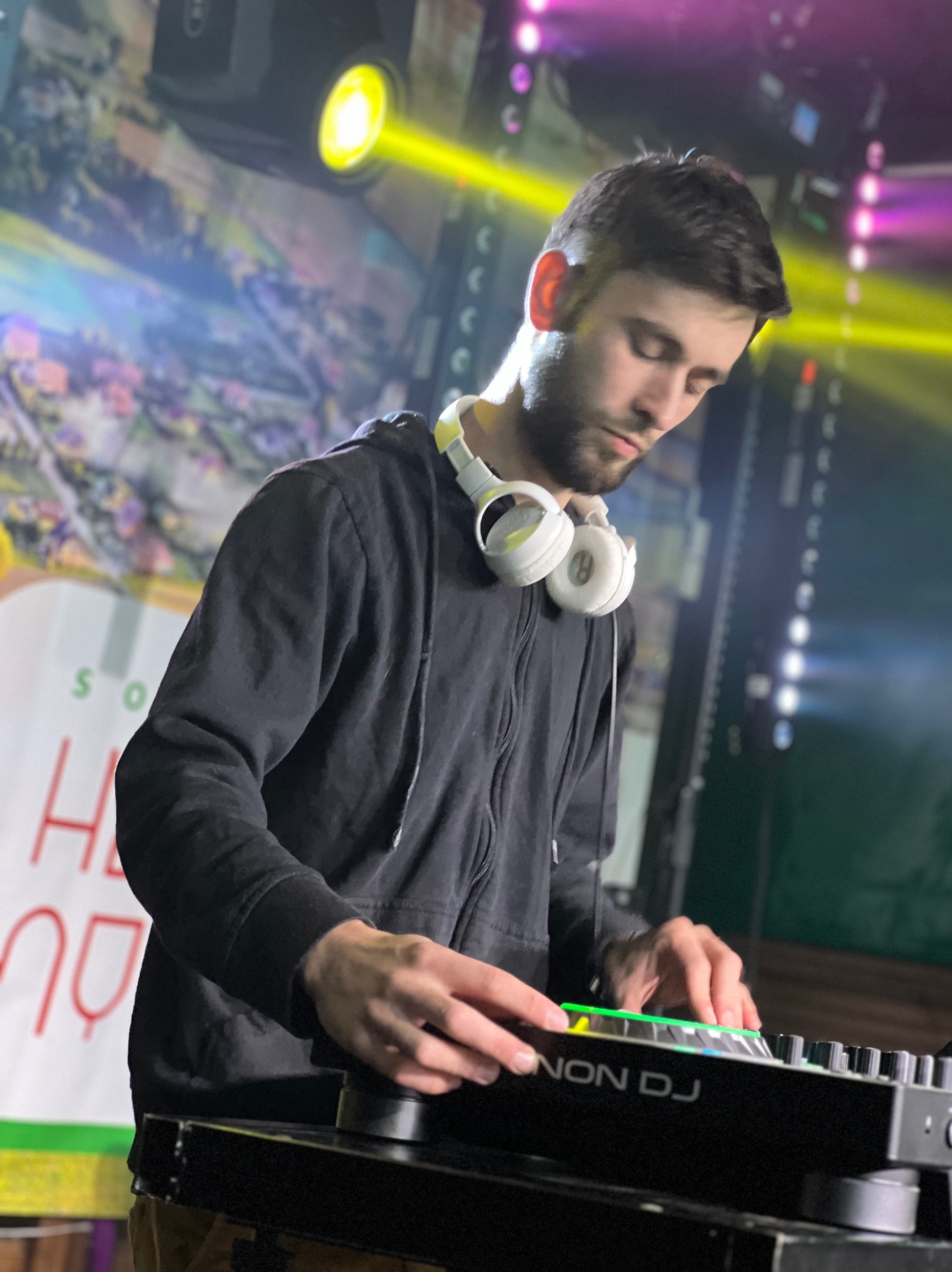 DJ w czarnej bluzie z kapturem, z białymi słuchawkami na szyi, miksuje muzykę na konsoli Denon DJ w oświetlonym klubie. Żółte i różowe światła tworzą imprezowy klimat.