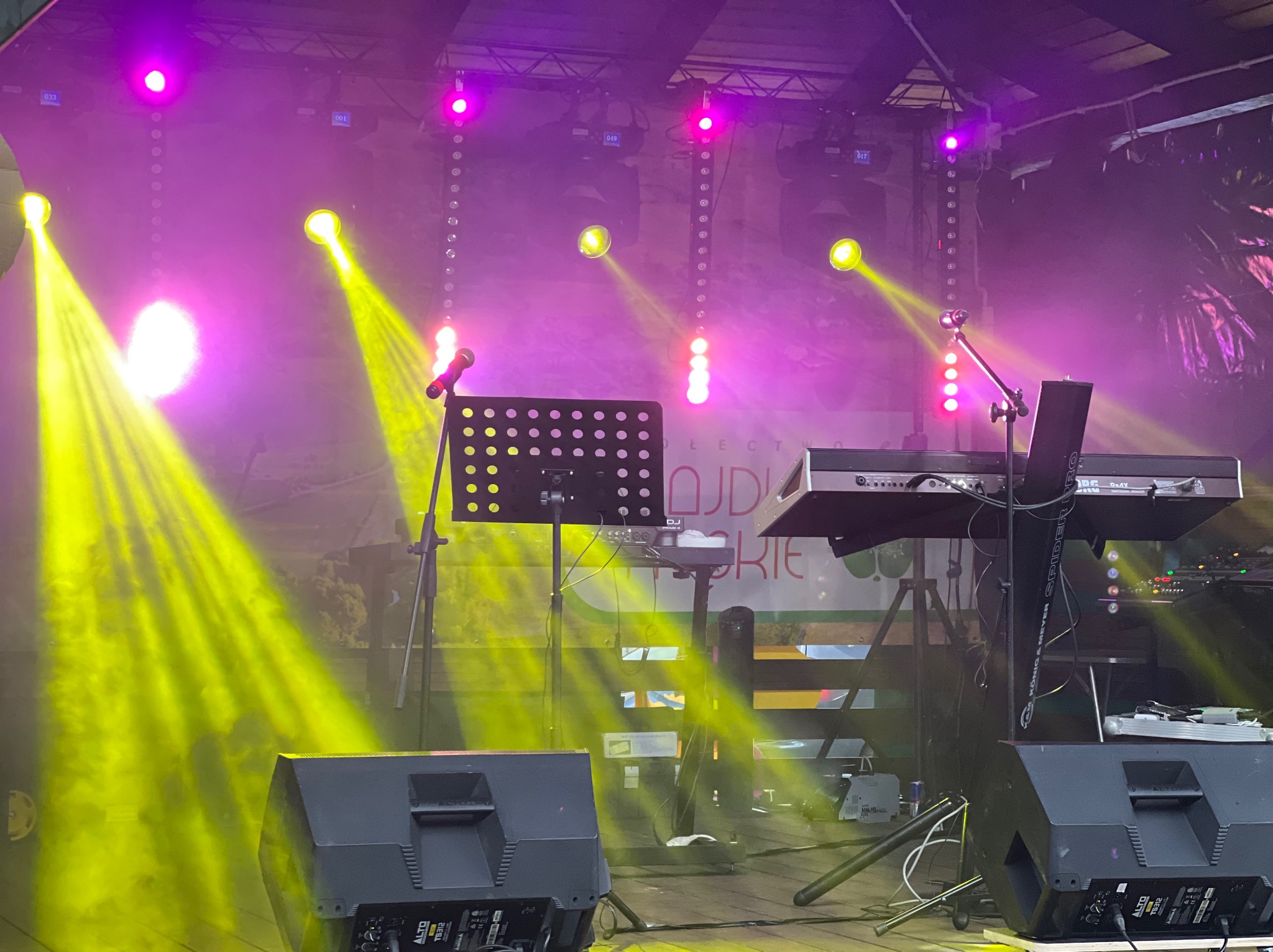 Scena z nagłośnieniem i oświetleniem w Maciejowicach, widoczne kolumny, keyboard i reflektory w odcieniach żółci i fioletu, gotowa do występu.