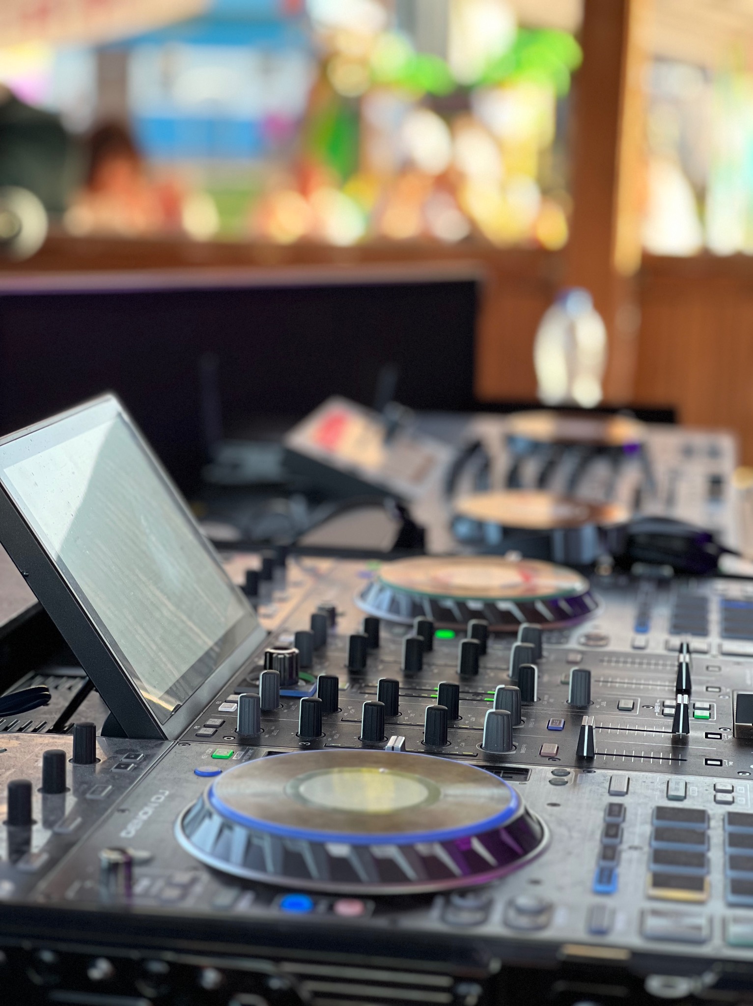 Profesjonalny kontroler DJ Denon z ekranem dotykowym, ustawiony na stole w plenerze, z rozmytym tłem imprezy. Widoczne pokrętła, suwaki i płyty gramofonowe.