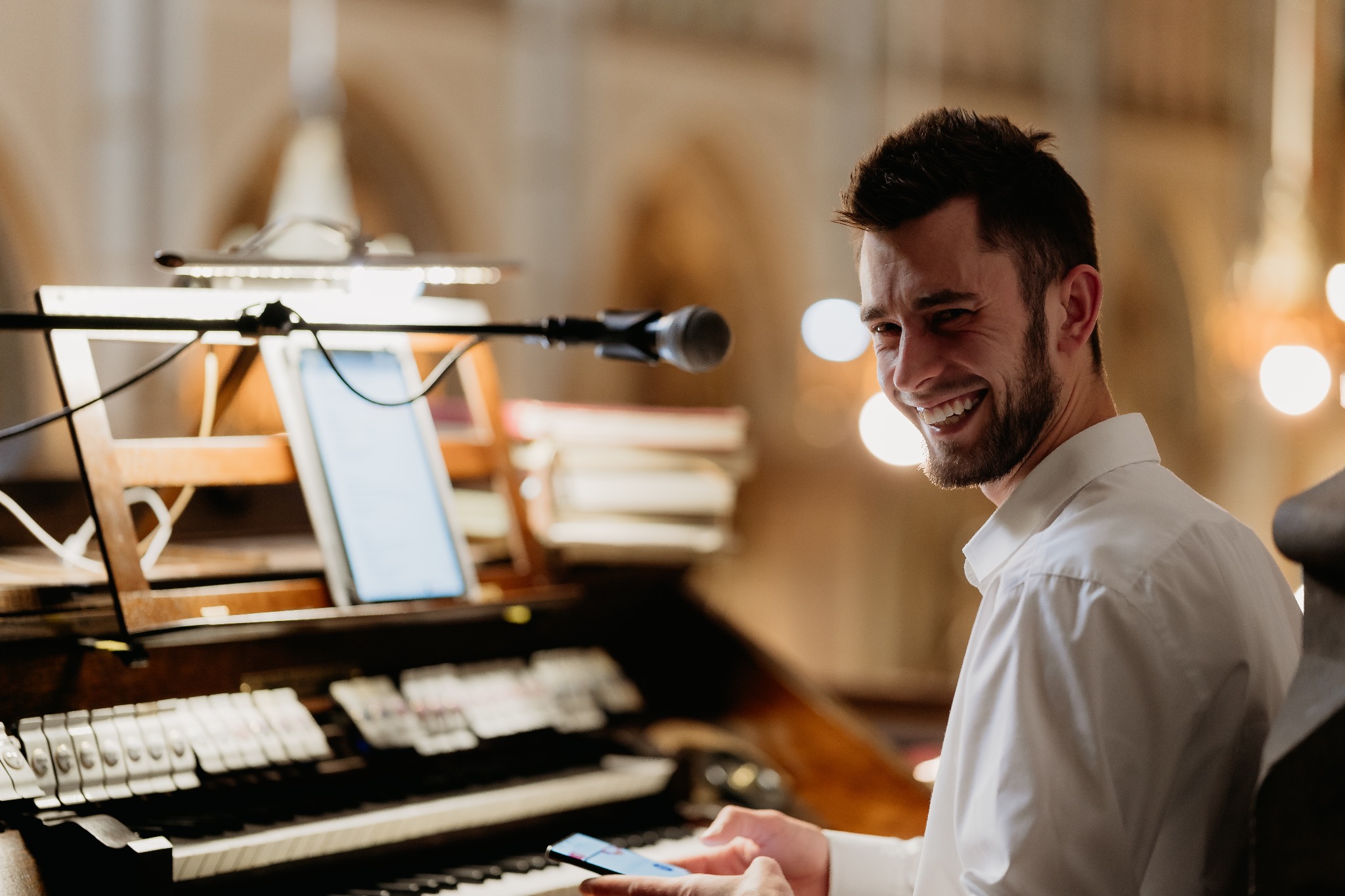 Uśmiechnięty mężczyzna w białej koszuli siedzi przy organach, trzymając smartfon. W tle widoczne oświetlenie i elementy architektoniczne wnętrza.