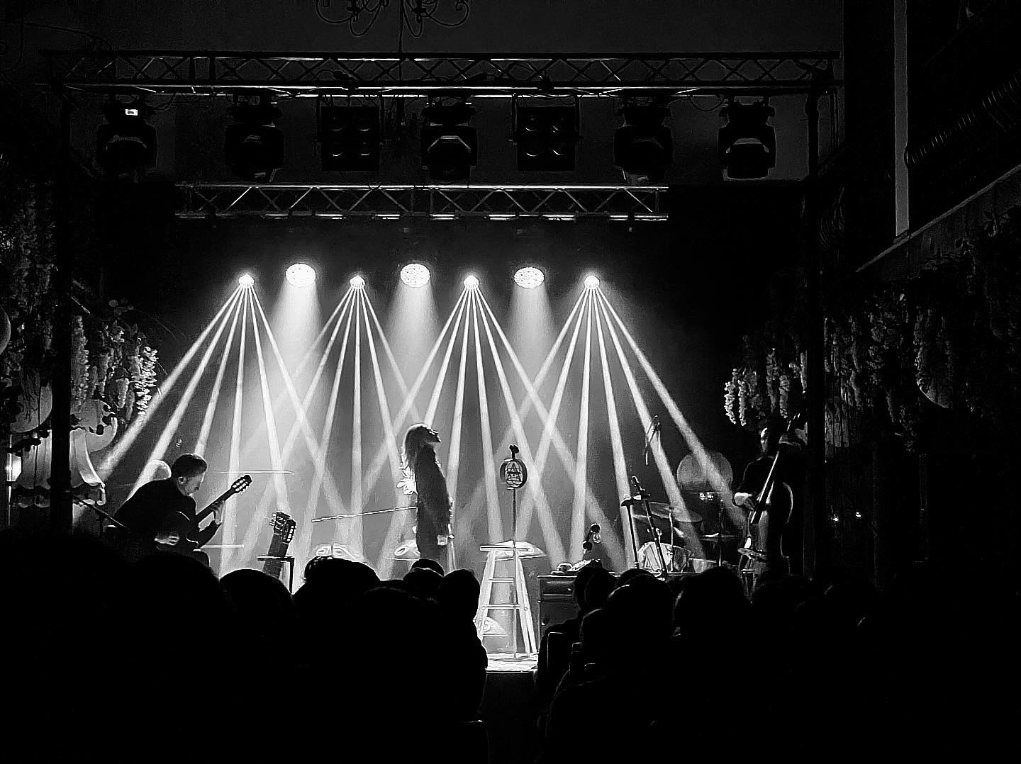 Czarno-białe zdjęcie zespołu muzycznego na scenie oświetlonej reflektorami. Wokalistka stoi przed mikrofonem, obok gitarzysta i kontrabasista. Widoczna publiczność.