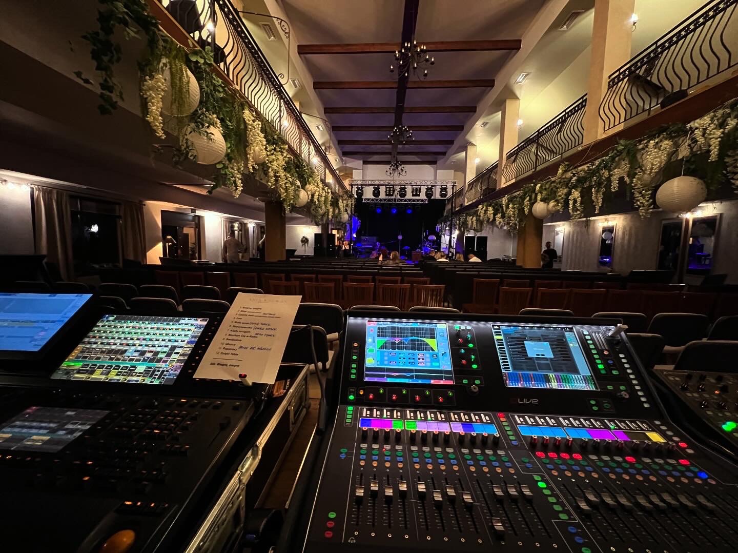 Konsoleta mikserska z kolorowymi suwakami na tle sali koncertowej z dekoracjami i sceną w oddali.