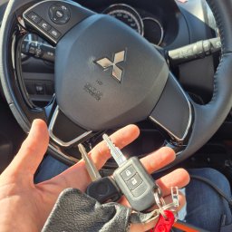 Womar Wojciechowski Mariusz - Kluczyki samochodowe Mitsubishi trzymane w dłoni na tle kierownicy, potencjalnie dorabianie kluczy lub awaryjne otwieranie auta.