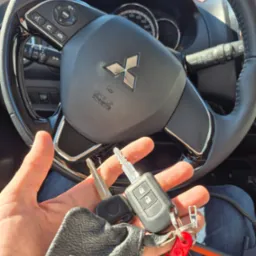 Kluczyki samochodowe Mitsubishi trzymane w dłoni na tle kierownicy, potencjalnie dorabianie kluczy lub awaryjne otwieranie auta.