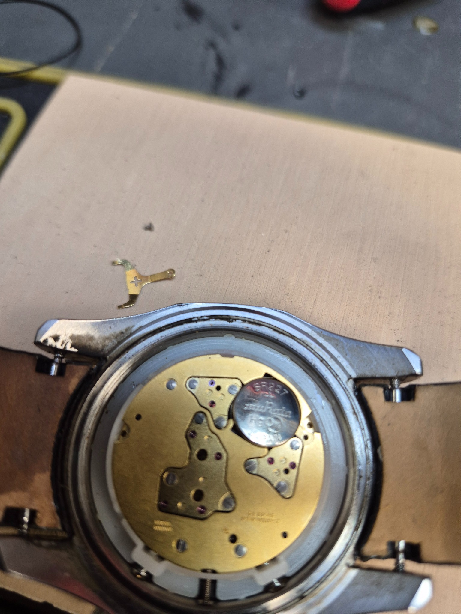 Otwarty zegarek z widocznym mechanizmem i baterią. Obok leży metalowy element mocujący. Detaliczna praca zegarmistrzowska.