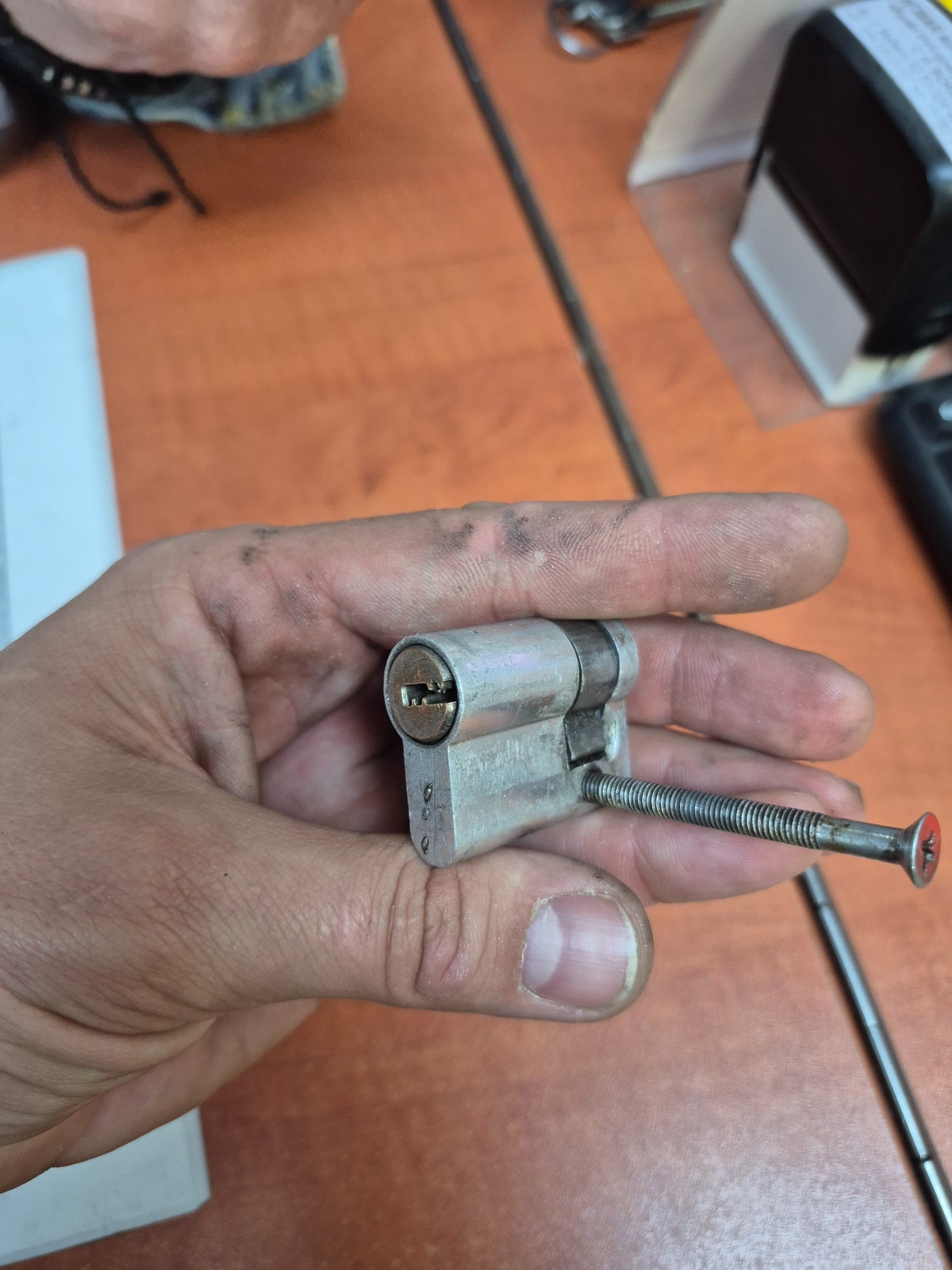 Otwarta wkładka zamka, trzymana w dłoni, ze śrubą wewnątrz, na tle biurka z przyborami. Precyzyjna praca przy zabezpieczeniach.