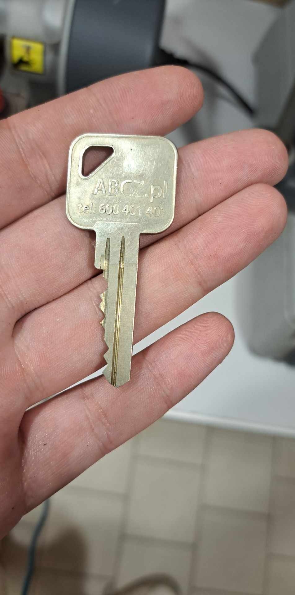 Detal: Nowy, srebrny klucz z wygrawerowanym adresem strony i numerem telefonu, leżący na otwartej dłoni. Widoczne unikalne nacięcia klucza.