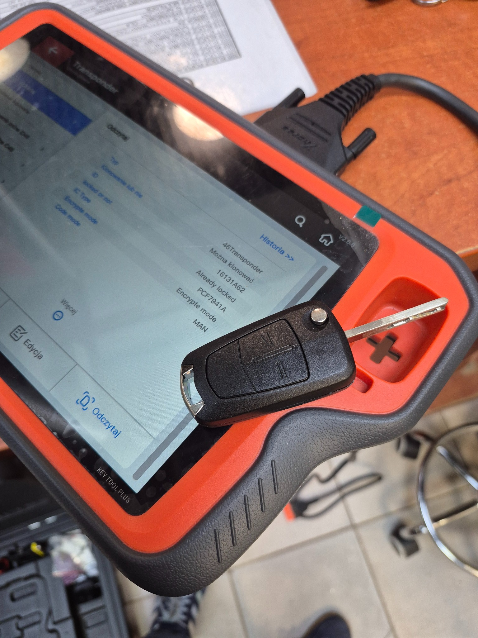 Kluczyk samochodowy leży na urządzeniu KEY TOOL PLUS z ekranem wyświetlającym dane transpondera. Urządzenie w pomarańczowo-czarnej obudowie, widoczne detale kluczyka.
