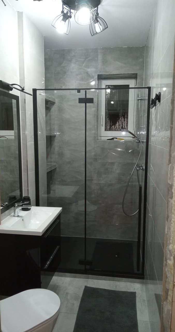 Nowoczesna łazienka z kabiną prysznicową typu walk-in, szare płytki imitujące beton, czarne akcenty i oświetlenie sufitowe. Minimalistyczny design.