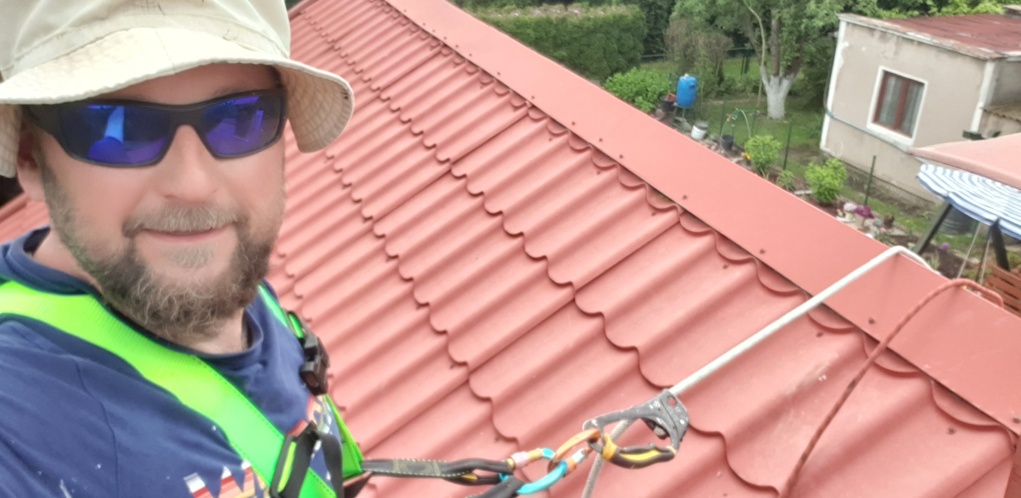 Pracownik w uprzęży na dachu z czerwonej dachówki falistej. Widoczny fragment ogrodu i budynek w tle. Bezpieczeństwo pracy na wysokości.