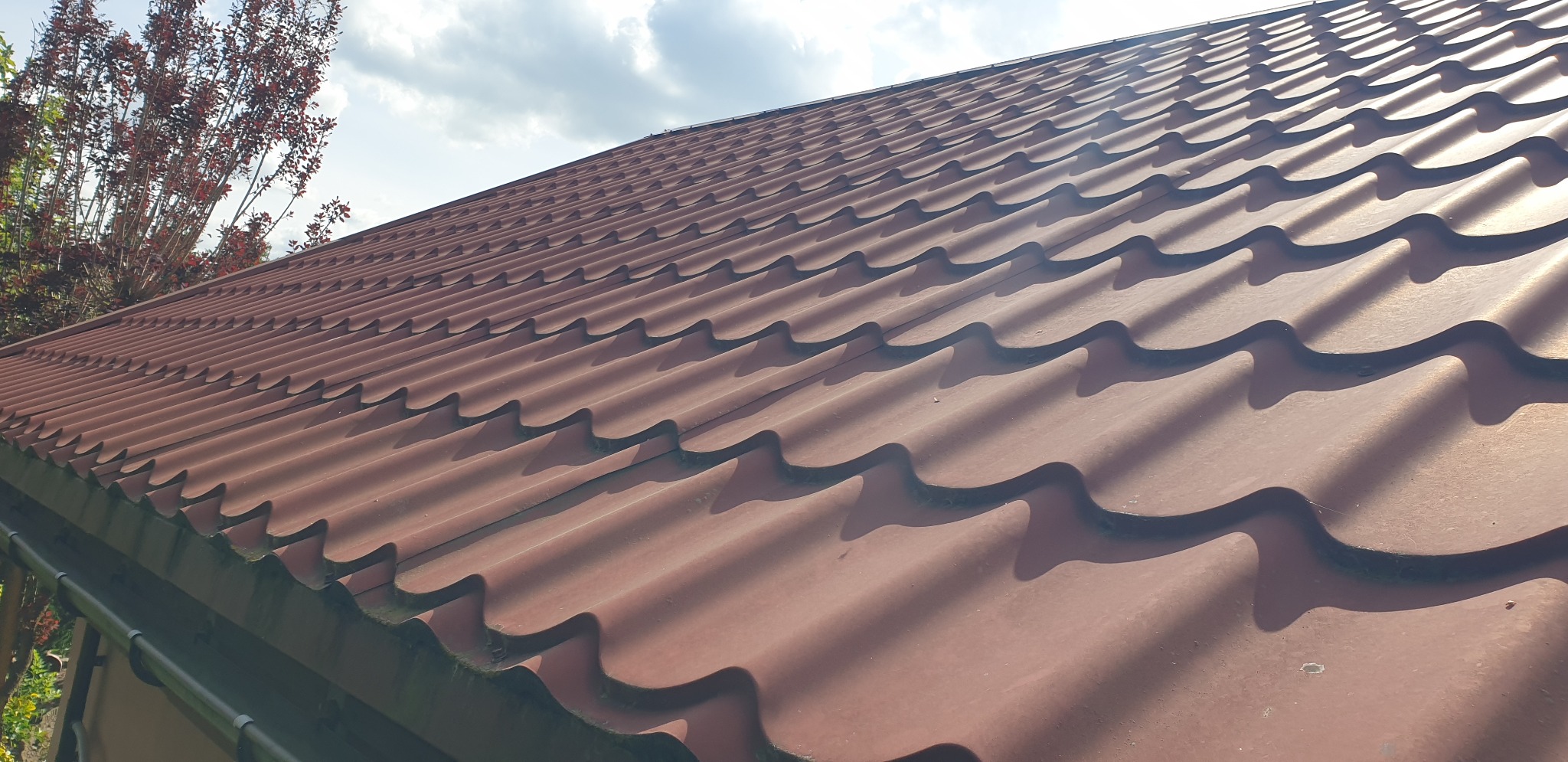 Czerwony dach z blachodachówki falistej, widoczny pod kątem. Na krawędzi dachu widoczne zabrudzenia. W tle drzewo i fragment błękitnego nieba.