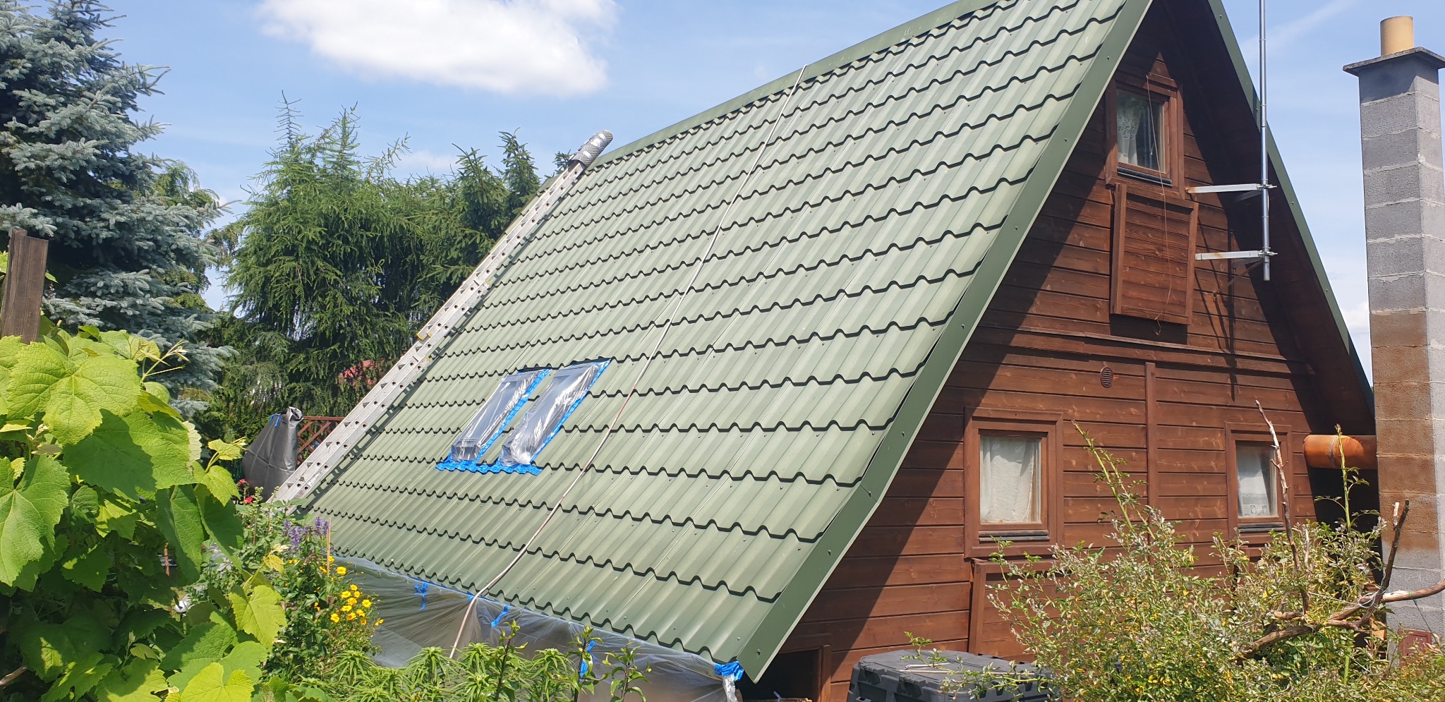 Dach domu z zieloną blachodachówką, zabezpieczony folią przy oknach dachowych. Przystawiona aluminiowa drabina. Widok na drewnianą elewację i komin.