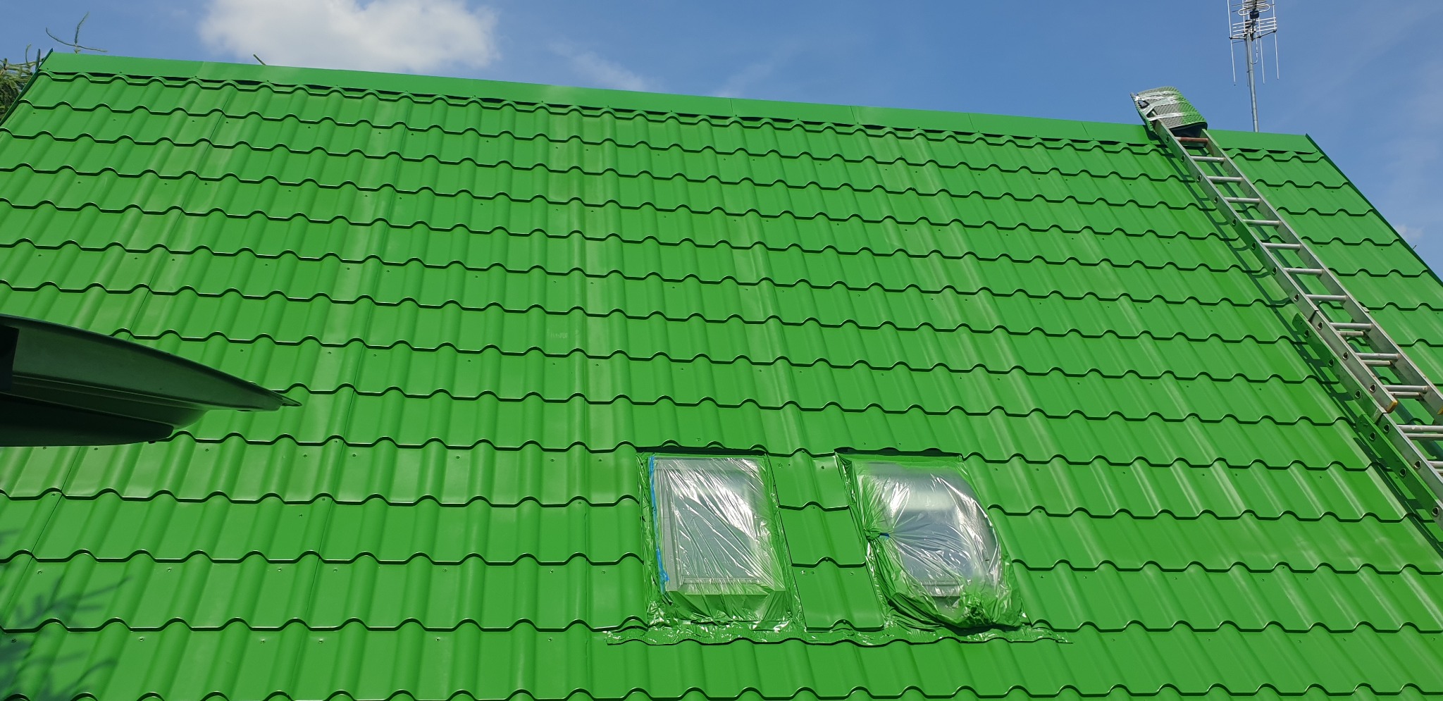 Jaskrawo zielony dach z blachodachówki po malowaniu; okna zabezpieczone folią, drabina oparta o krawędź dachu, antena TV w tle na tle błękitnego nieba.