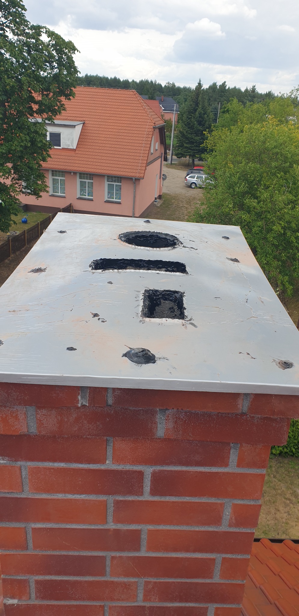 Czapka kominowa z otworami w trakcie wymiany, widok z góry na ceglany komin z szarą, metalową czapką. W tle budynek mieszkalny i drzewa.