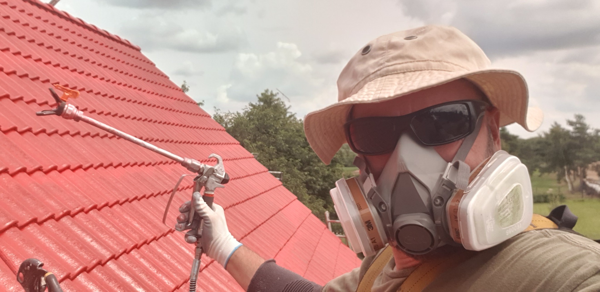 Pracownik w masce maluje czerwony dach pistoletem natryskowym. Widoczne zabezpieczenia osobiste i fragment krajobrazu w tle. Kąt ujęcia z dołu.