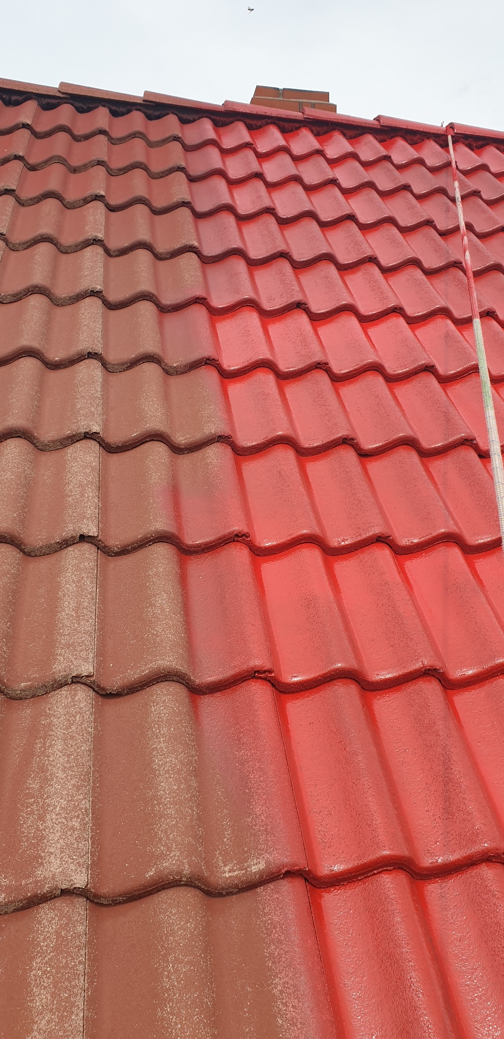 Dachówka ceramiczna: połowa dachu pomalowana na czerwono, druga w odcieniu brązu. Widoczny fragment komina i lina. Prezentacja efektu malowania dachu.