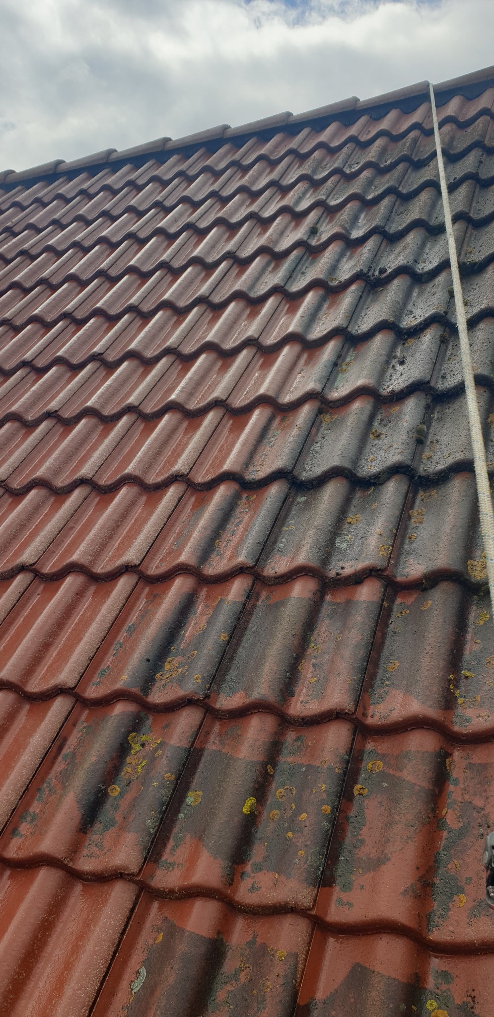Częściowo umyty dach z czerwonej dachówki, widoczne różnice w czystości. Liny zabezpieczające w prawym górnym rogu kadru. Pochmurne niebo w tle.