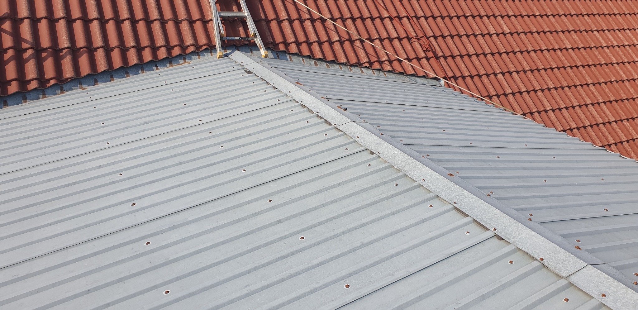 Szary dach z blachy trapezowej, widoczne mocowania, fragment dachu pokrytego czerwoną dachówką w tle, drabina oparta o dach. Ujęcie z góry.