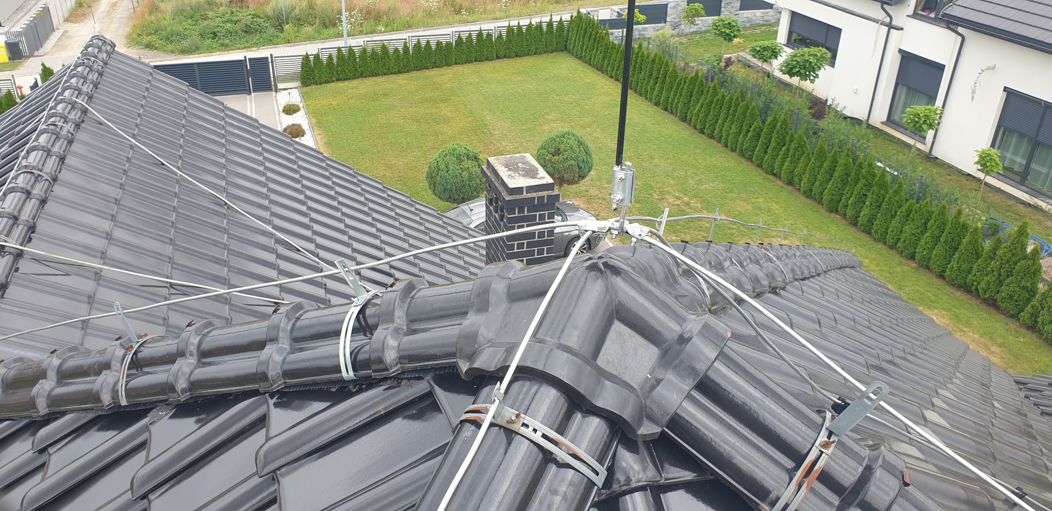Czarny dach z dachówki ceramicznej po instalacji odgromowej, widoczne przewody i uchwyty mocujące, w tle trawnik i dom z szarą stolarką okienną.