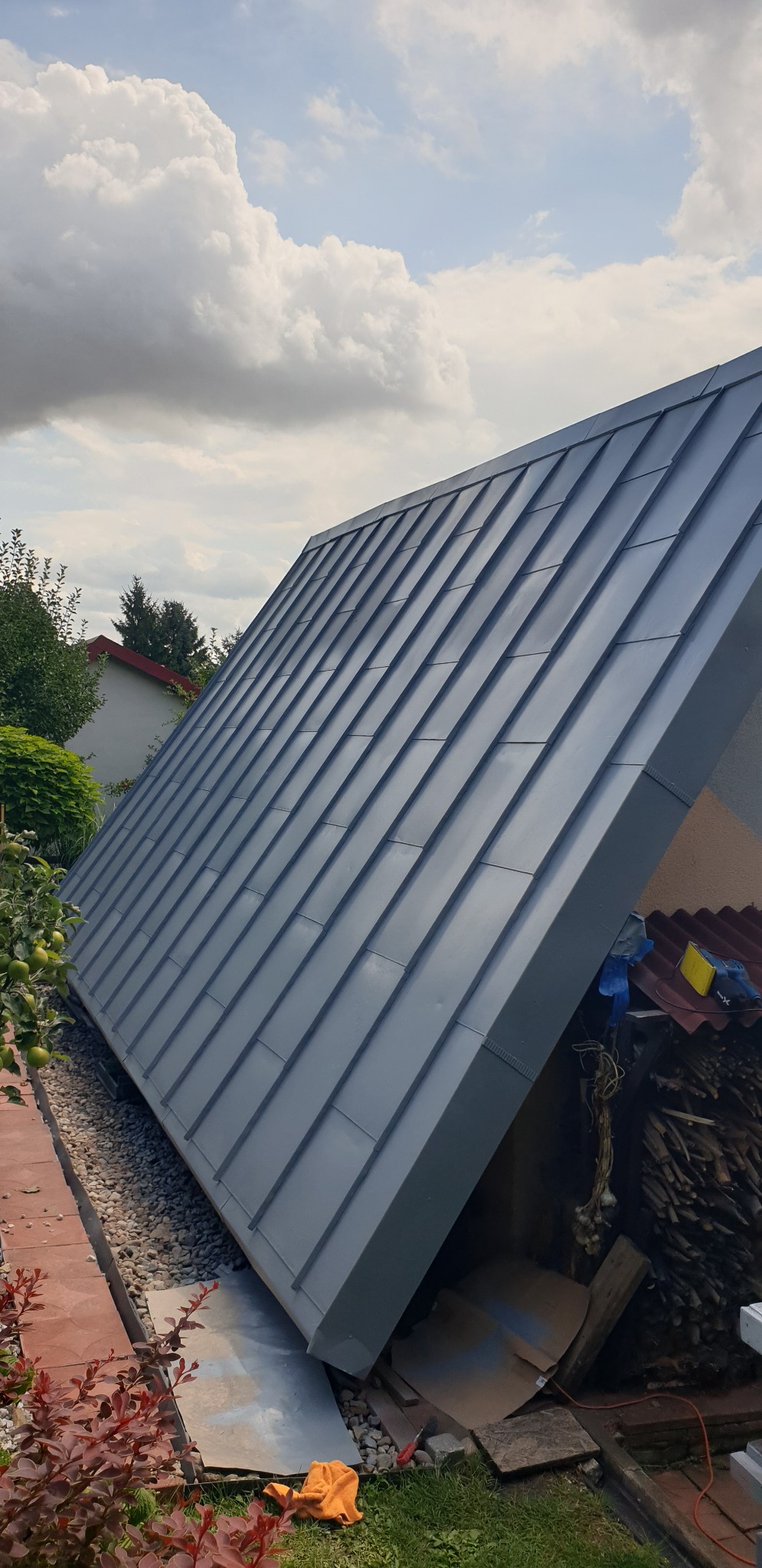 Szary, metalowy dach po malowaniu, widoczny z boku, na tle zieleni i pochmurnego nieba. Dach pokryty panelami na rąbek stojący. Widoczne narzędzia.