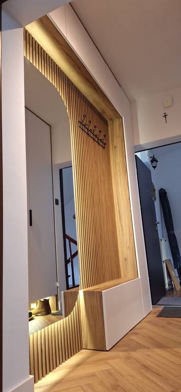 Nowoczesna zabudowa przedpokoju z drewnianymi lamelami, siedziskiem i wieszakiem, uzupełniona o lustro. Widoczna podłoga ułożona w jodełkę.