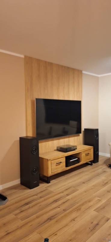 Drewniana zabudowa TV z szafką RTV i głośnikami. Widok na salon z parkietem. Minimalistyczny design, ciepłe oświetlenie. Realizacja.