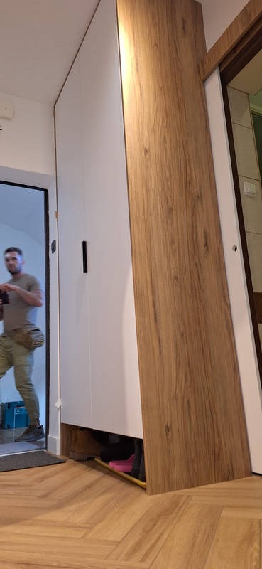 Biało-drewniana szafa wnękowa z czarnym uchwytem, widoczna z perspektywy podłogi, z elementami wyposażenia widocznymi pod spodem i fragmentem osoby w tle.