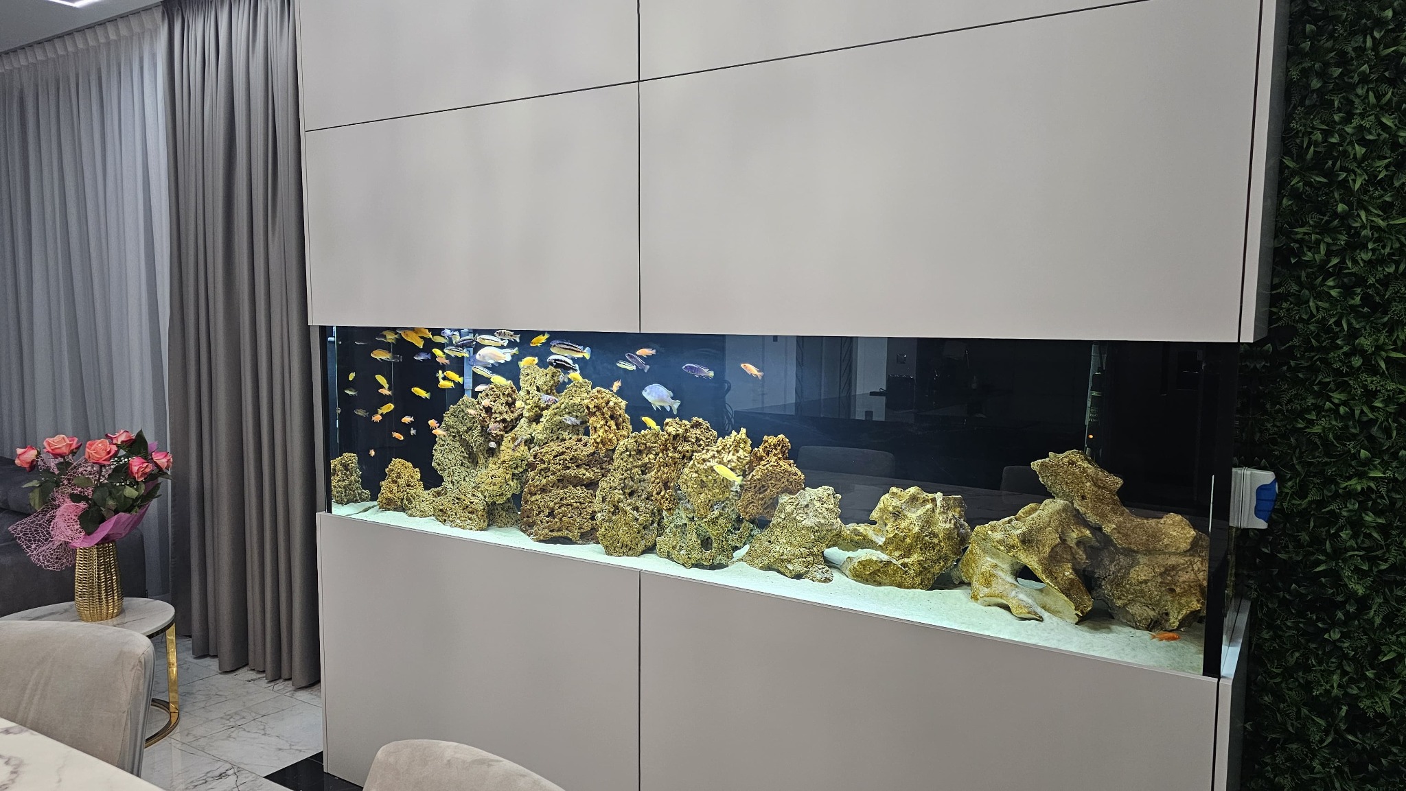 Wnętrze z akwarium w zabudowie meblowej, widoczne ryby i dekoracje skalne, z boku ściana z roślinnością, obok bukiet kwiatów. Całość w nowoczesnym stylu.
