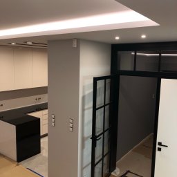 VLAD BUD - Nowoczesna kuchnia z czarnym blatem i wyspą, widoczna przez otwarte, przeszklone drzwi w czarnej ramie. Podwieszany sufit z oświetleniem LED. Minimalistyczny design.