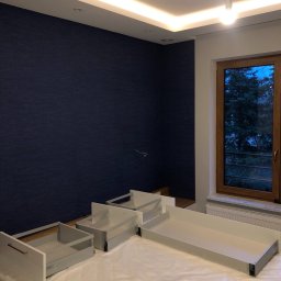 VLAD BUD - Pokój z ciemnoniebieską ścianą i rozłożonymi elementami mebli, prawdopodobnie szufladami, przygotowanymi do montażu. Widoczne okno i podwieszany sufit z oświetleniem.