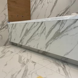 VLAD BUD - Wykończona łazienka z płytkami imitującymi marmur na podłodze, ścianach i zabudowie wanny. Widoczne fragmenty instalacji elektrycznej i drewnianej ściany.