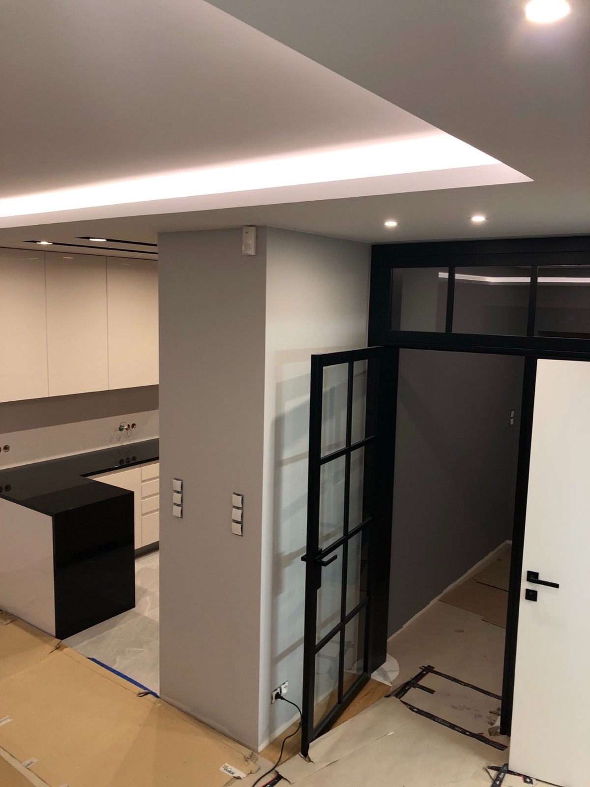 Nowoczesna kuchnia z czarnym blatem i wyspą, widoczna przez otwarte, przeszklone drzwi w czarnej ramie. Podwieszany sufit z oświetleniem LED. Minimalistyczny design.