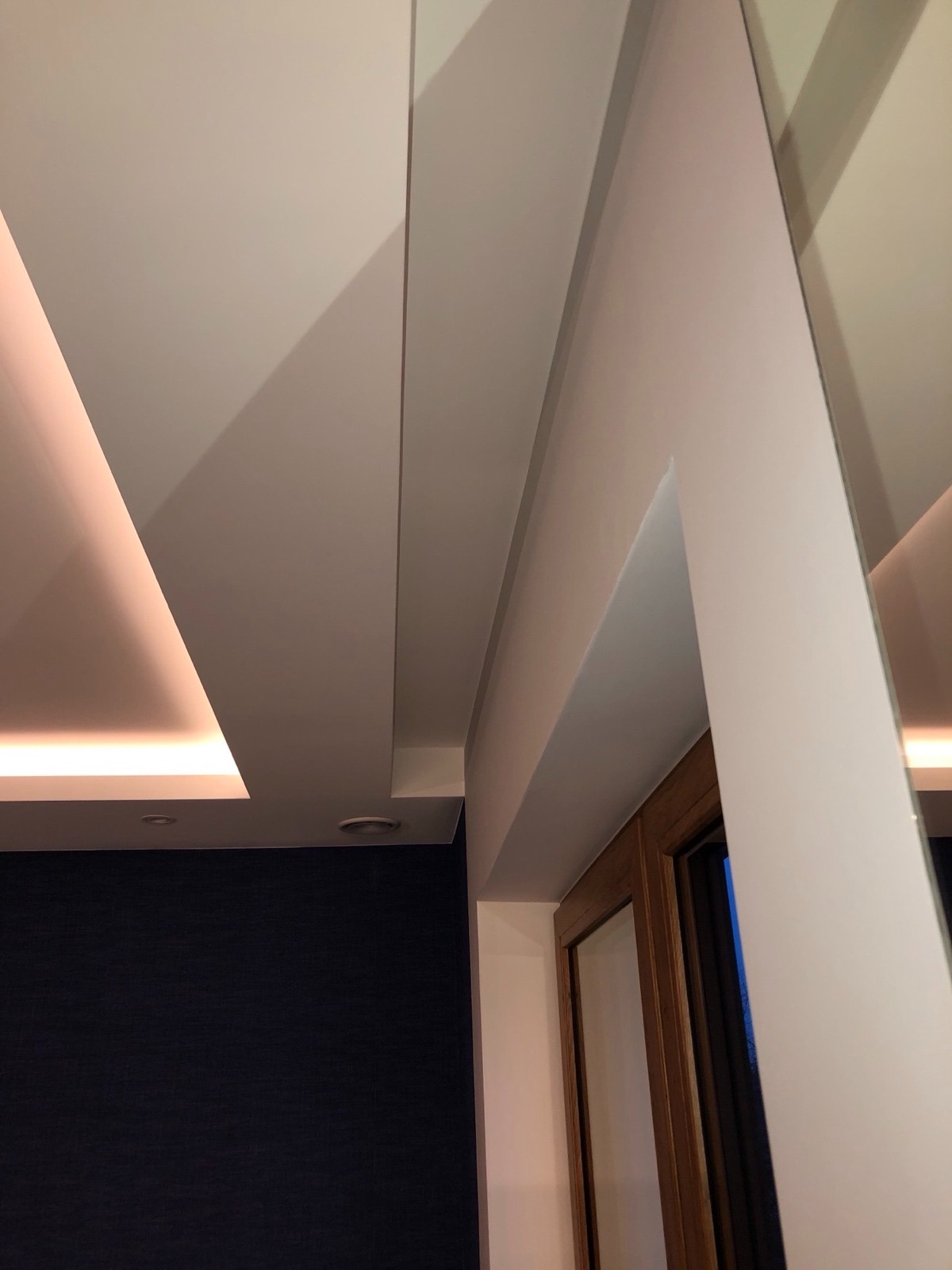 Nowoczesny sufit podwieszany z oświetleniem LED i zabudową nad ościeżnicą drzwiową. Ciemna ściana w tle. Nietypowy kąt ujęcia.