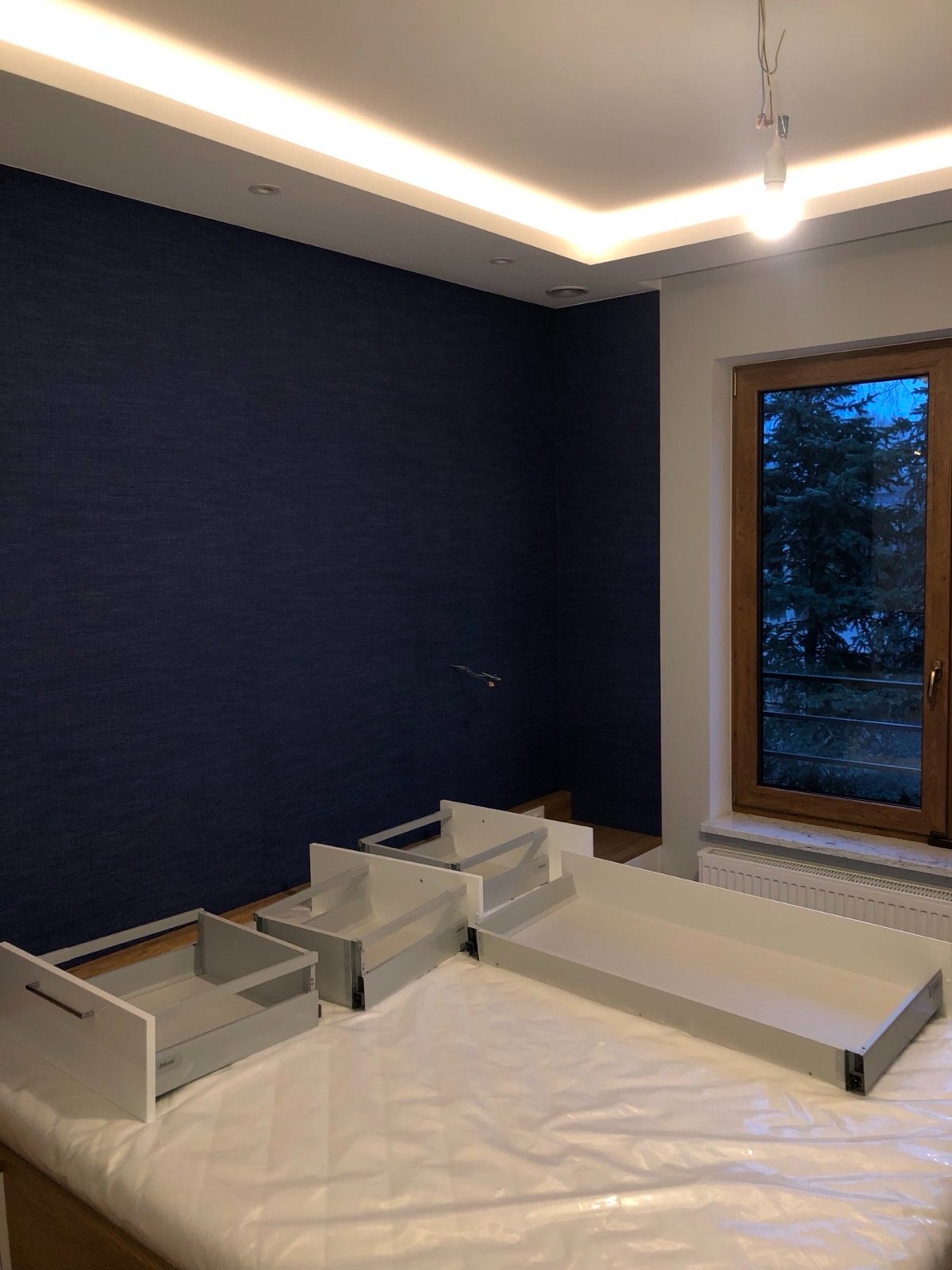 Pokój z ciemnoniebieską ścianą i rozłożonymi elementami mebli, prawdopodobnie szufladami, przygotowanymi do montażu. Widoczne okno i podwieszany sufit z oświetleniem.