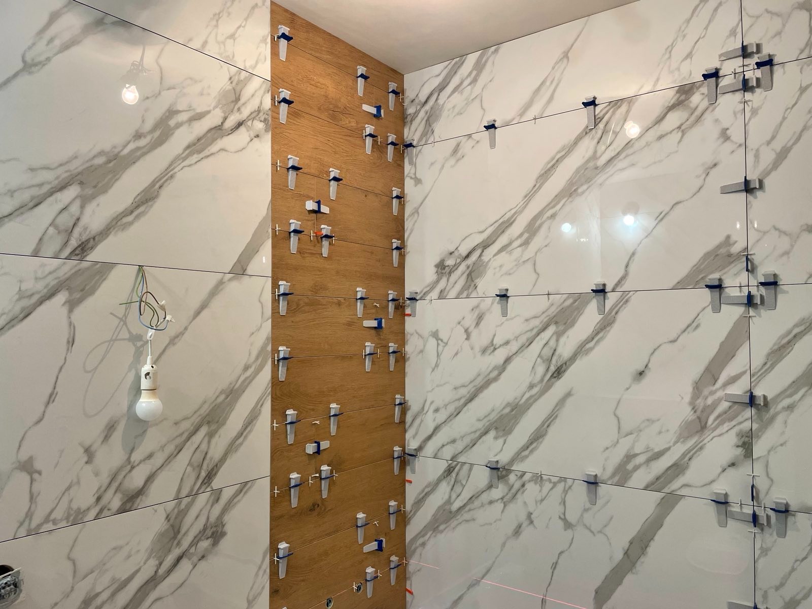Ułożone płytki imitujące marmur i drewno na ścianach łazienki, z systemem poziomowania płytek, oświetleniem i wiązką lasera. Prace glazurnicze w toku.