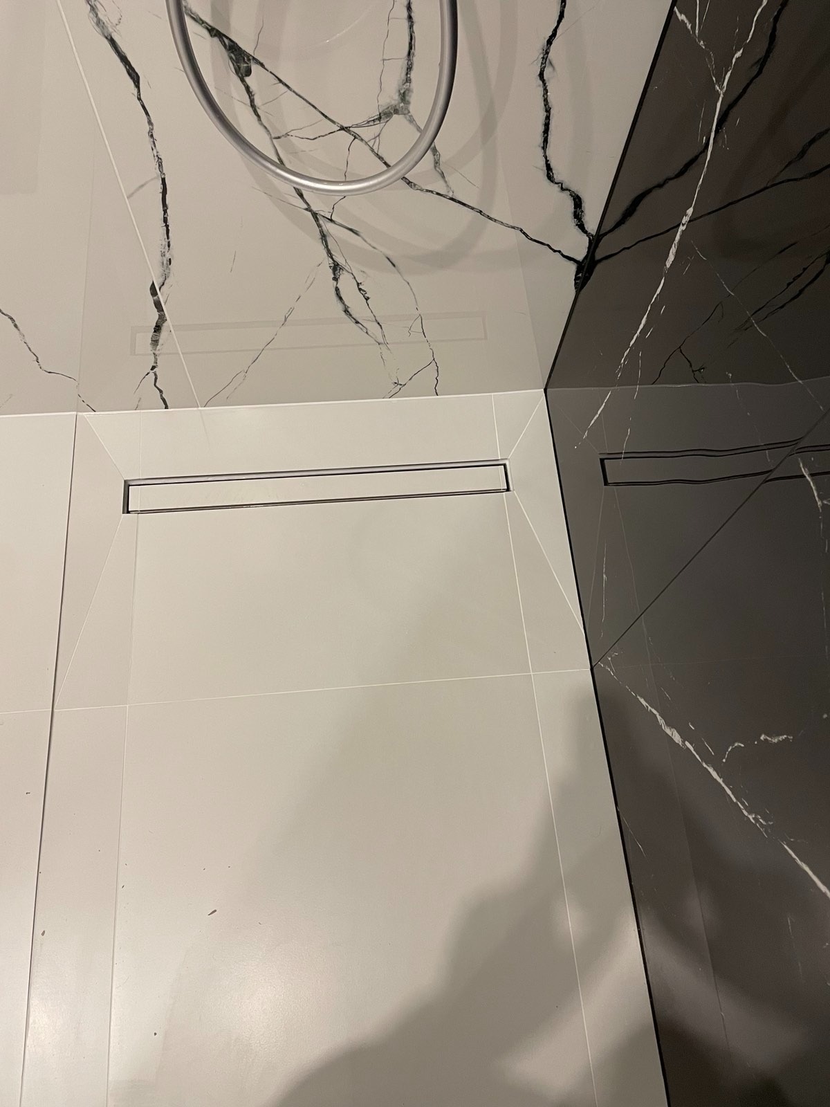 Nowoczesna łazienka z odpływem liniowym i marmurowymi płytkami w odcieniach bieli i czerni. Widok z góry na podłogę i fragment ściany z deszczownicą.