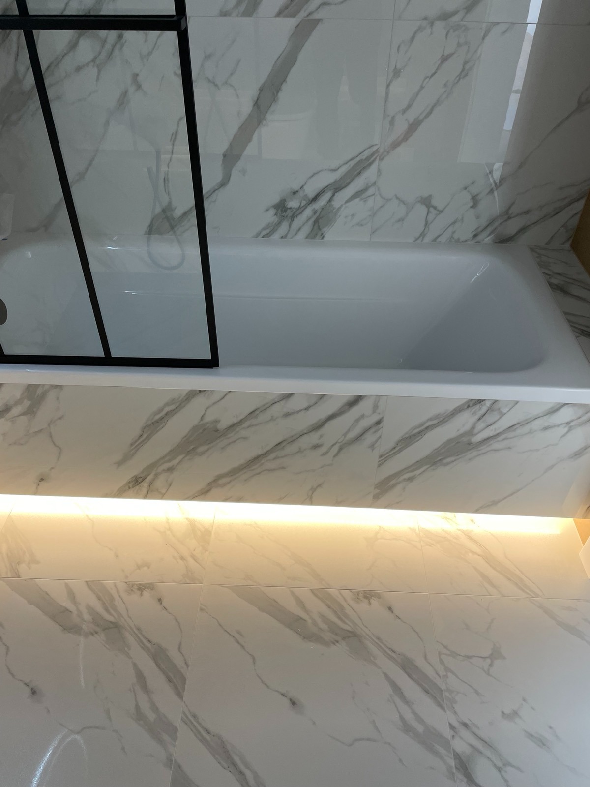 Biała wanna z czarną szybą kabiny, marmurowe płytki na ścianach i podłodze, podświetlenie LED pod wanną. Minimalistyczny design łazienki.