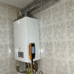 Biały piec gazowy De Dietrich na tle płytek, z podłączonym analizatorem spalin Testo 300, rura odprowadzająca spaliny u góry. Widoczne przyłącza gazowe i elektryczne.