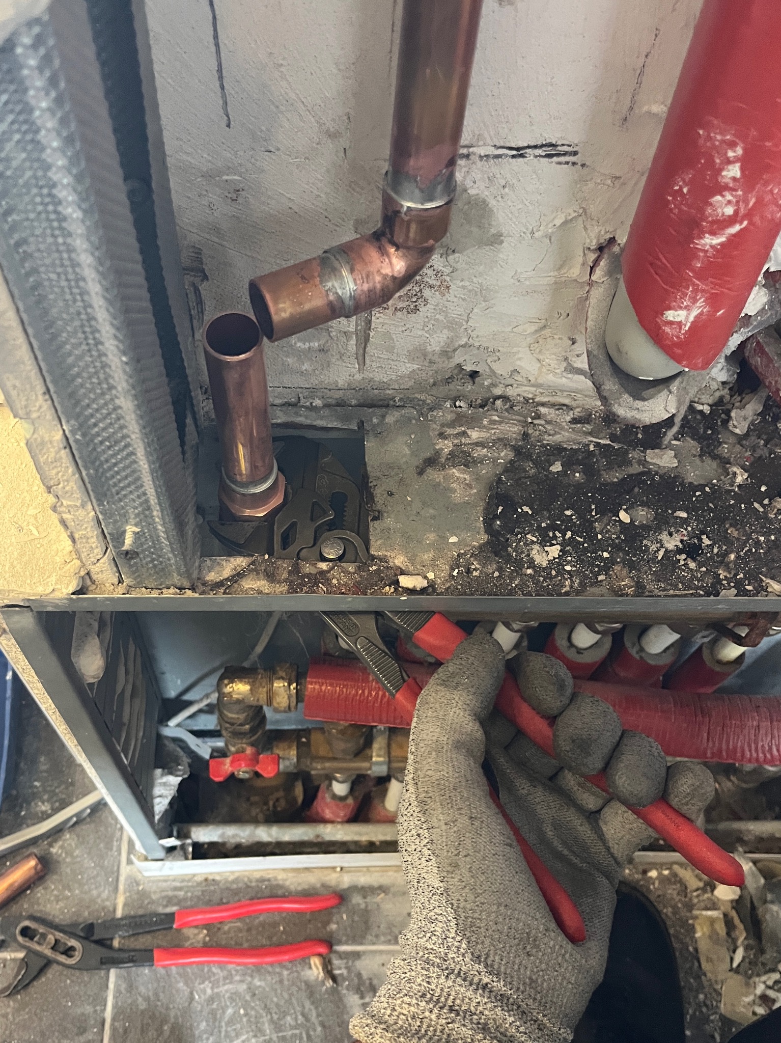 Instalacja rur miedzianych w kotłowni, widoczne ręce w rękawicach trzymające narzędzia, elementy instalacji grzewczej i fragment ściany z instalacjami.