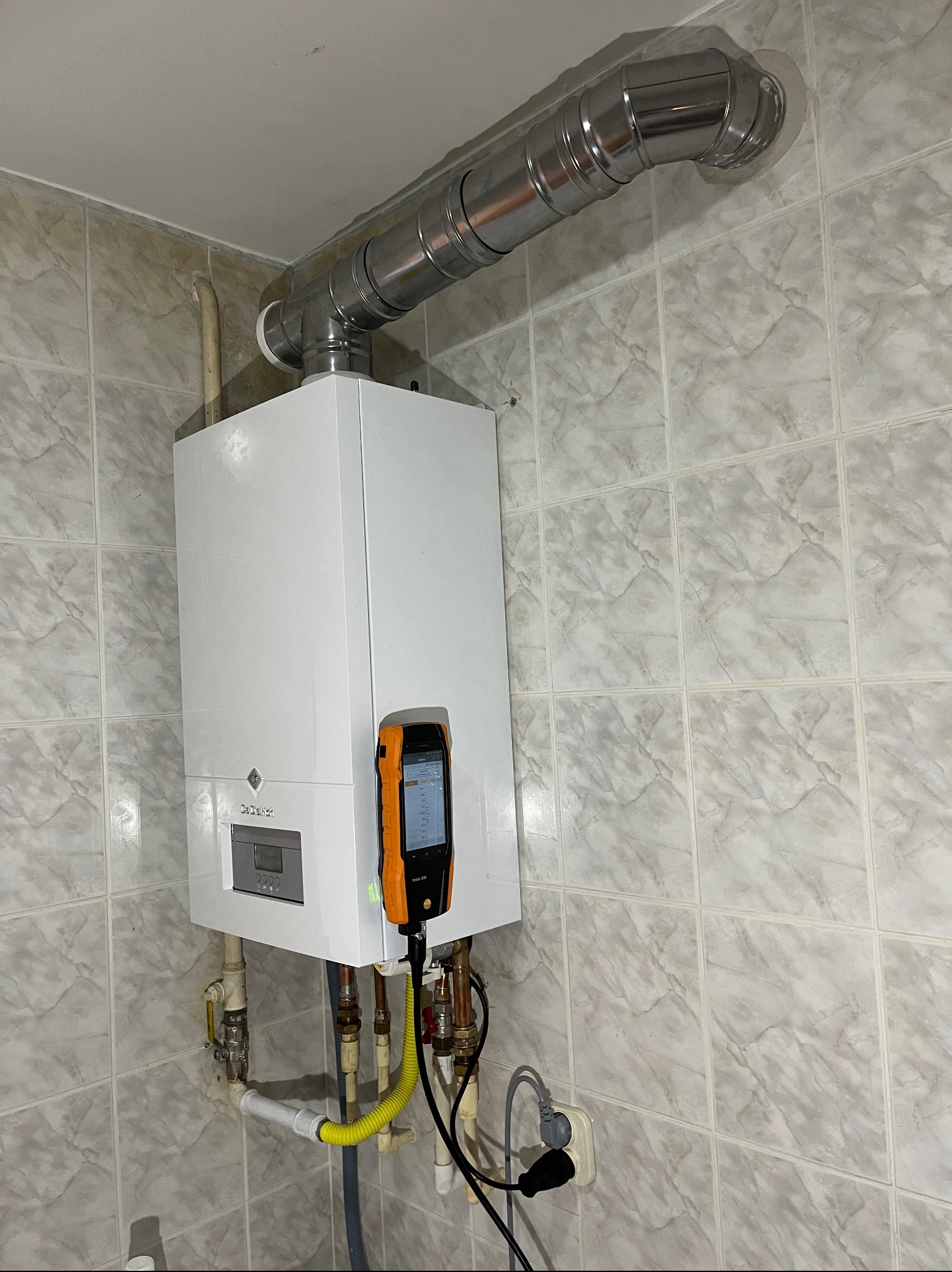 Biały piec gazowy De Dietrich na tle płytek, z podłączonym analizatorem spalin Testo 300, rura odprowadzająca spaliny u góry. Widoczne przyłącza gazowe i elektryczne.