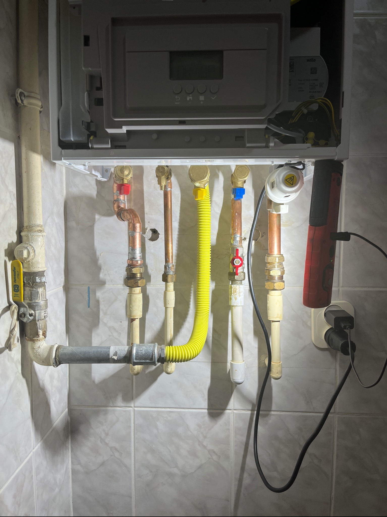 Podłączenie kotła gazowego z widocznymi rurami miedzianymi, zaworami i żółtym przewodem gazowym na tle kafelkowanej ściany. Widoczna lampa warsztatowa.