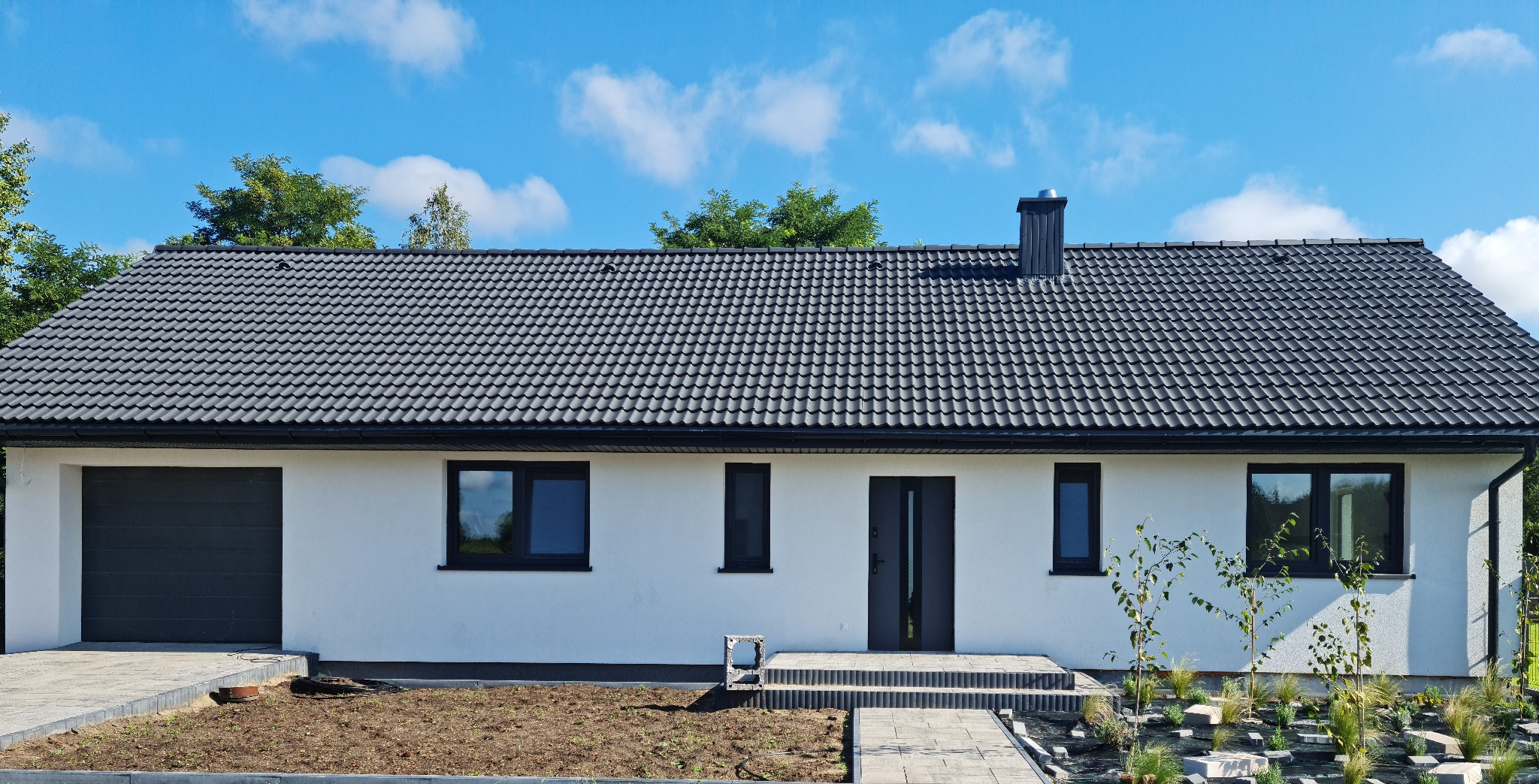 Parterowy dom z ciemną dachówką i grafitową stolarką okienną, na tle błękitnego nieba z delikatnymi obłokami. Widoczny fragment zagospodarowanego terenu.