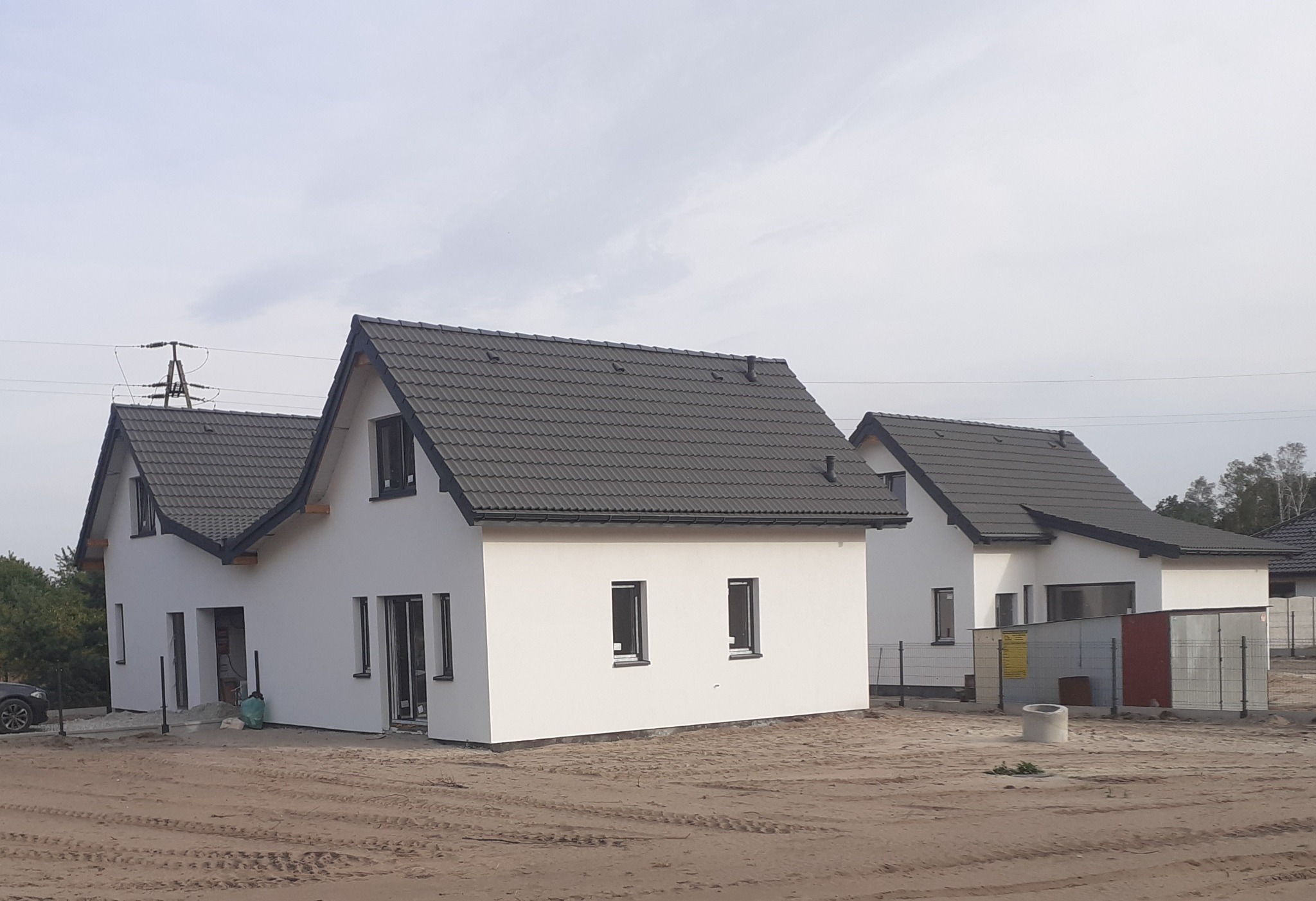 Nowoczesne, białe domy jednorodzinne z ciemnymi dachami na działce w trakcie budowy. Widoczne elementy infrastruktury i ogrodzenie tymczasowe. Ujęcie z poziomu gruntu.