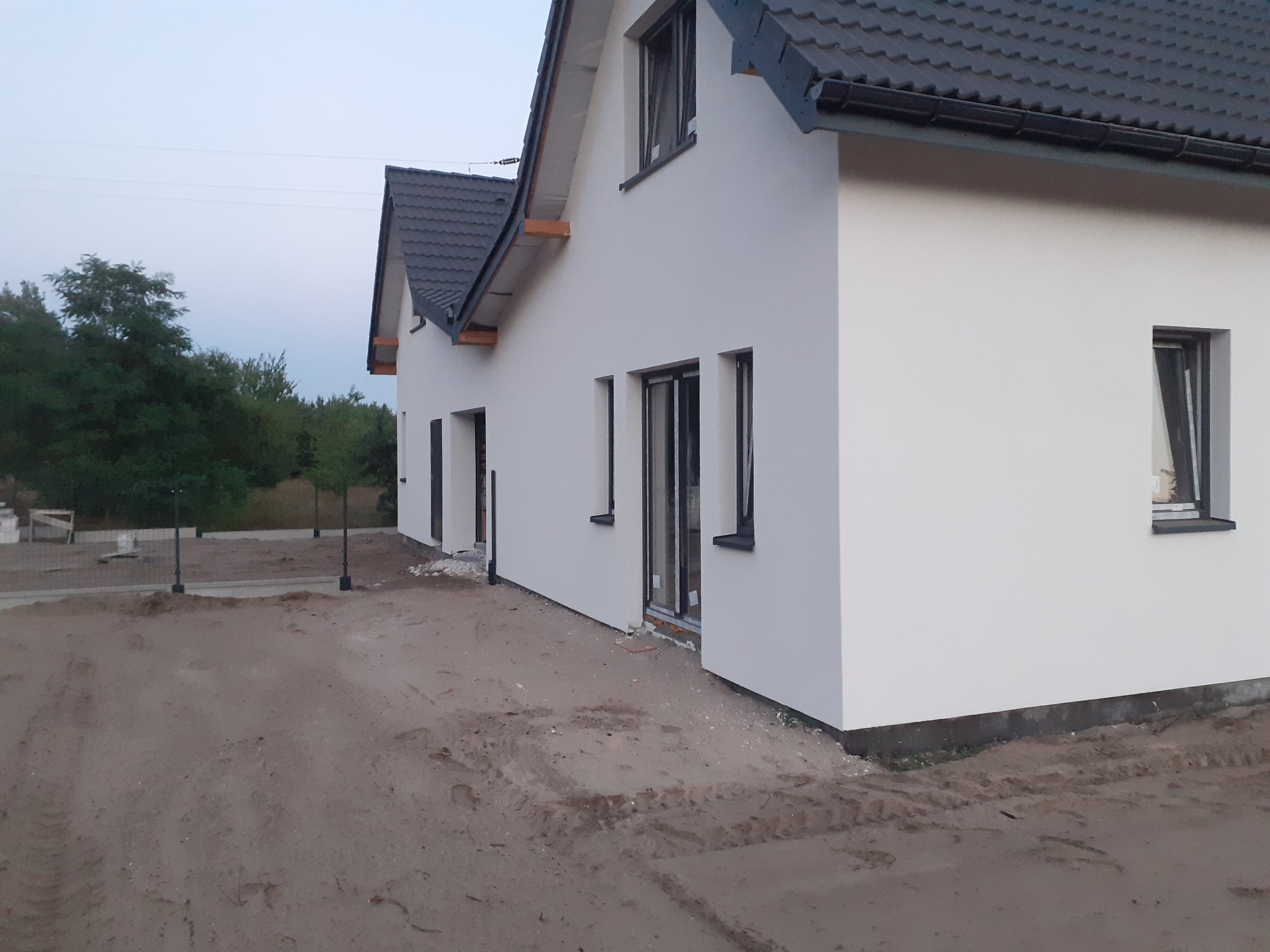 Nowoczesny, biały dom z ciemną dachówką, widok z boku. Stolarka okienna w kolorze grafitowym. Teren wokół domu w trakcie zagospodarowania.