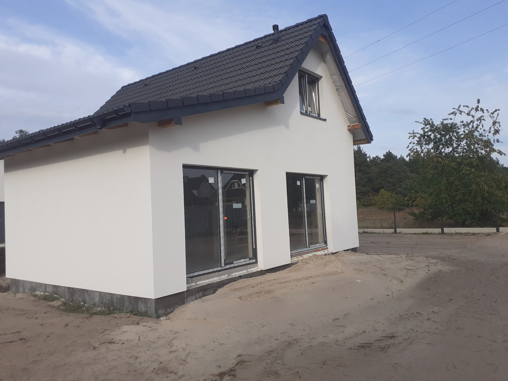 Nowoczesny dom z białymi ścianami i ciemnym dachem z dachówki, widoczne duże okna i przygotowany teren wokół budynku. Detal: brak rynien.