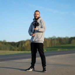 DIAXOR DESIGN - projektowanie animacji i realizacja zleceń video - Młody kamerzysta z brodą, w bluzie z kapturem, trzyma profesjonalną kamerę ze stabilizatorem na tle błękitnego nieba i zielonych drzew. Ujęcie z dołu, pełna sylwetka.