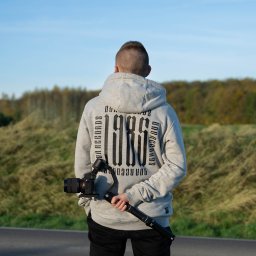 DIAXOR DESIGN - projektowanie animacji i realizacja zleceń video - Mężczyzna tyłem w bluzie z kapturem trzymający stabilizator kamery z aparatem na tle krajobrazu. Ujęcie od tyłu, skupione na sprzęcie i otoczeniu.