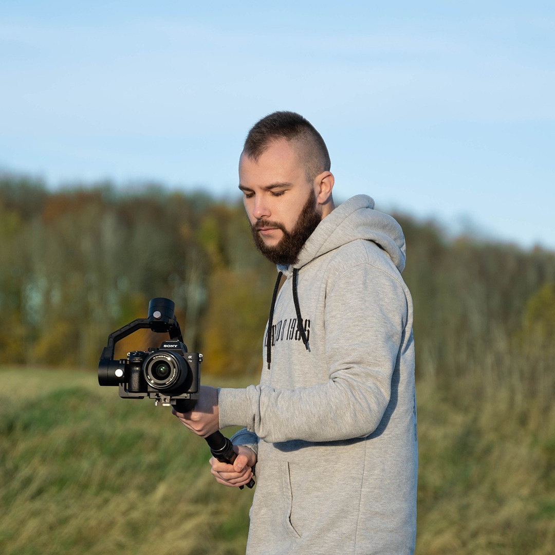 Młody mężczyzna z brodą w szarej bluzie z kapturem obsługuje profesjonalny aparat Sony z gimbalem na tle łąki i drzew. Dobre oświetlenie.