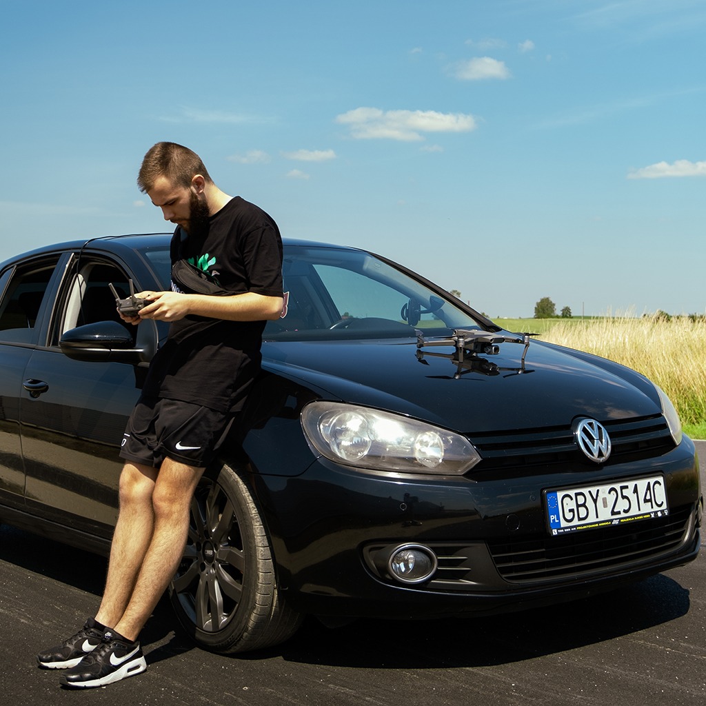 Młody mężczyzna oparty o czarny samochód, trzymający kontroler drona. Na masce auta leży dron. W tle pole i błękitne niebo z chmurami.