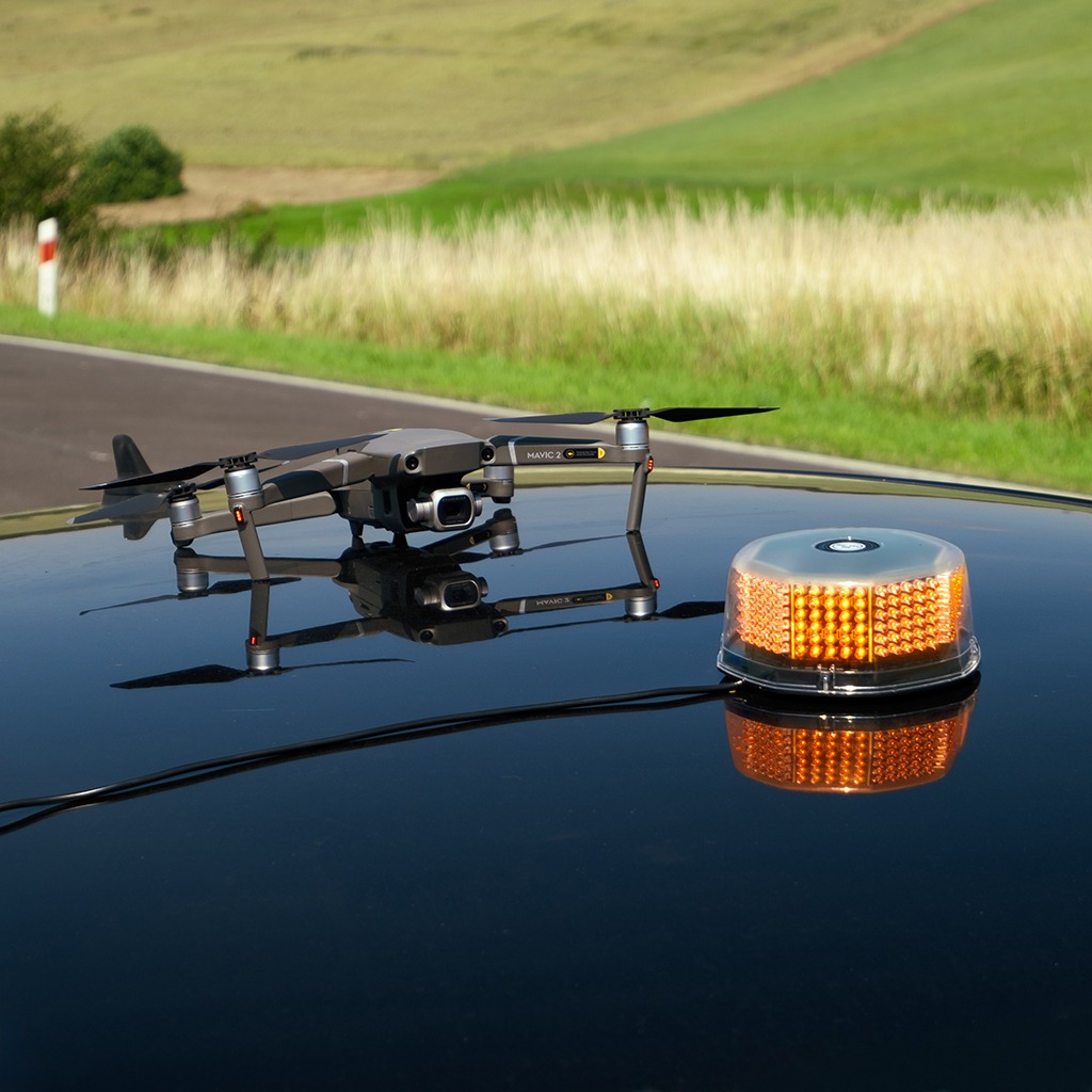 Dron Mavic 2 z kamerą na dachu czarnego samochodu obok pomarańczowego światła ostrzegawczego; w tle droga i zielone wzgórza w słoneczny dzień.